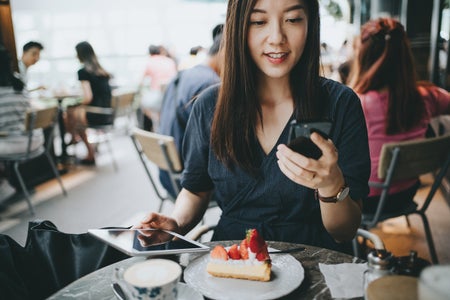 Busy young woman using smartphone while working on digital tablet in an outdoor cafe