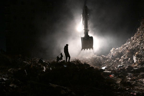 Silhouette of person next to bulldozer sifting through debris from collapsed building.