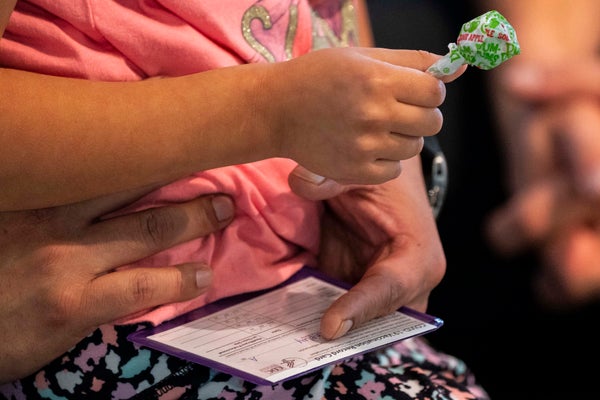 Child's hand grasps a lollipop while the parent holds a vaccination card on their lap.