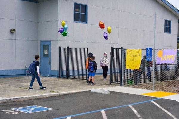 Young, masked, students walk into school entrance decorated with balloons.