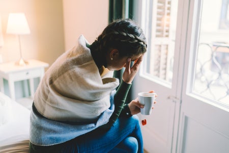 A woman wrapped in a blanket with her hand on her face while holding a cup of tea.