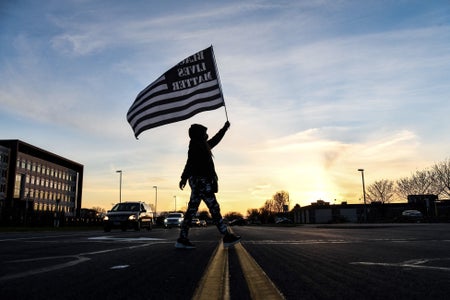 A demonstrator marches crosses tthe street, holding a Black Lives Matter flag against the sunset.