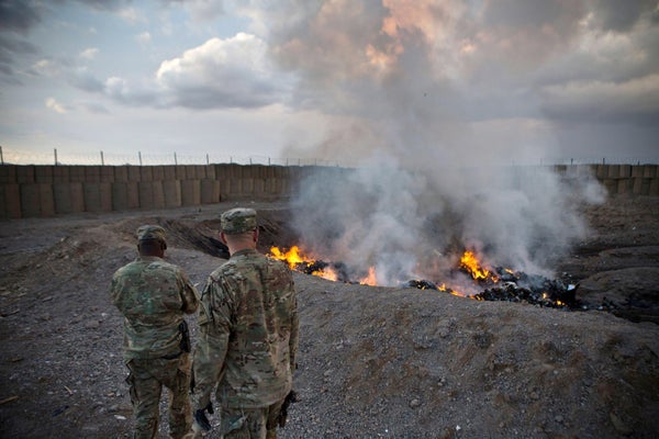 Soldiers watch smoke billow from a burn pit in the ground.