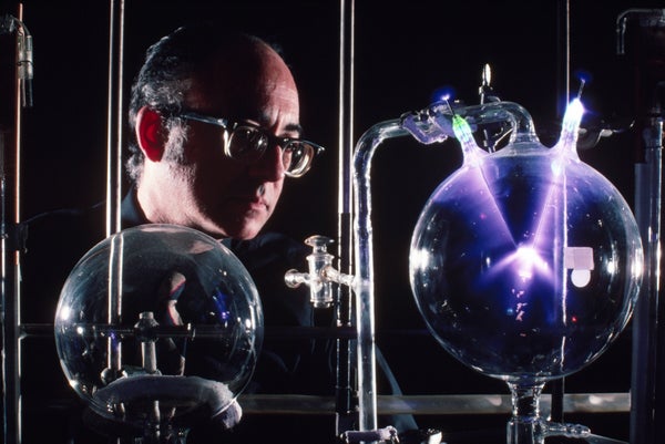 A man looks into a laboratory glass flask that's glowing with purple light.