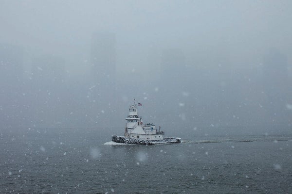 Snowy scene of a river with a tugboat and city obscured by snow in the background.