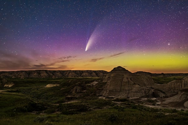 A comet is seen in the purple and yellow sky above badlands.