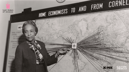 In a black and white photo a woman stands in front of a world map pointing to a swarm of push pins with strings tied to them that spread outward from a single point on the map