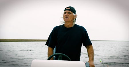 A man with a baseball cap turned backward stands at the controls onboard a small skiff on the ocean.