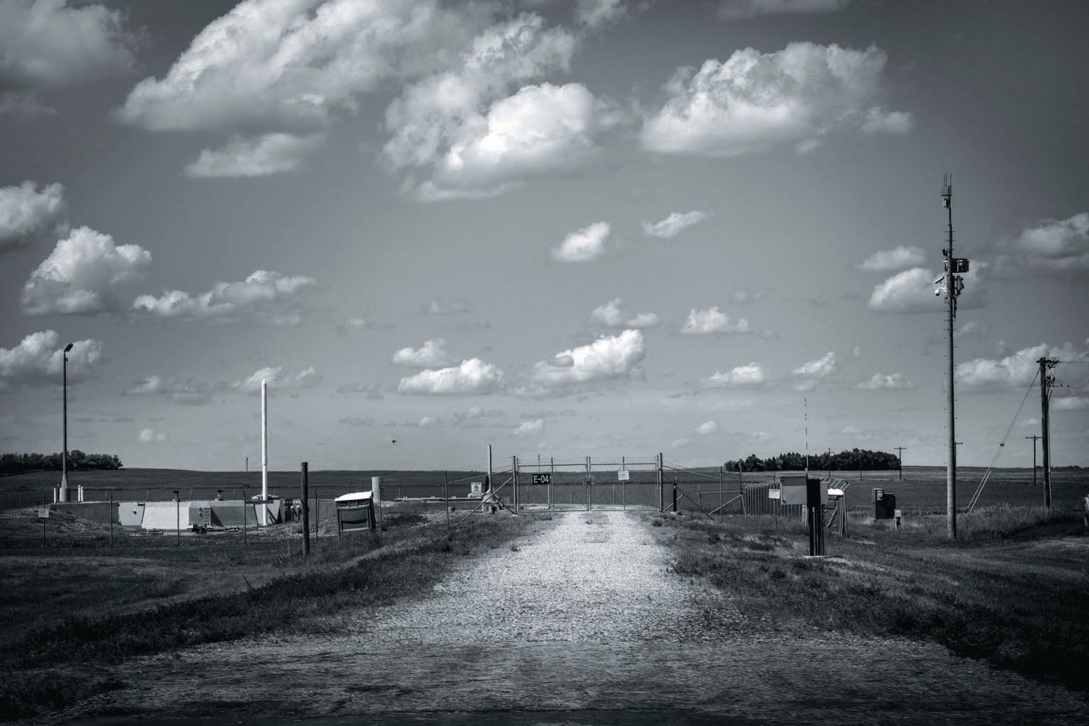 Black and white photograph showing a remote area with dirt road and metal gate.