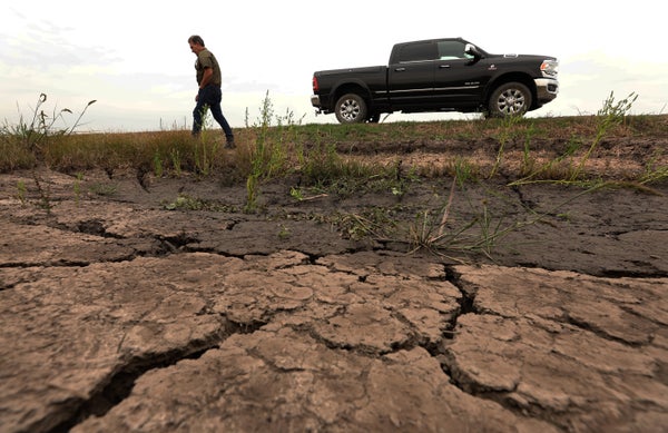 Cracked earth during a severe drought that would normally be underwater is seen in the foreground, in the background a farmer walks near his parked truck as he surveys the area
