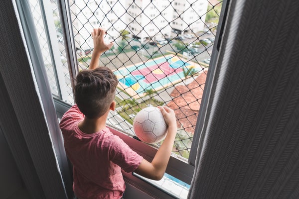 A boy holding a ball, seen from behind, gazes out of a window.n