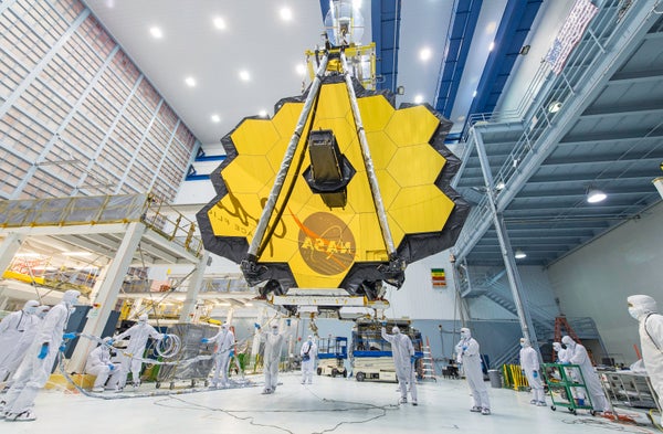 Workers in white bodysuits surround suspended James Webb Space Telescope
