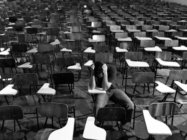 Black and white photo of large room full of empty desks and one lone student sitting with their head in their hands
