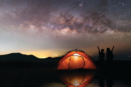 A lit tent outdoors with two people pointing up at the stars.