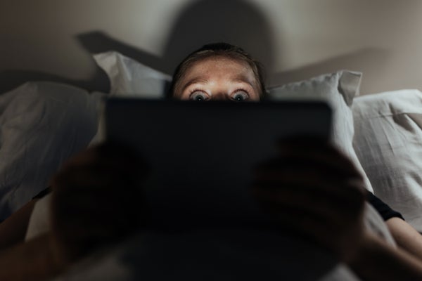 Scared woman in bed peeking up behind her laptop with scared eyes.