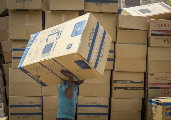 A man carries a boxed A/C unit, with stack of other A/Cs seen in background