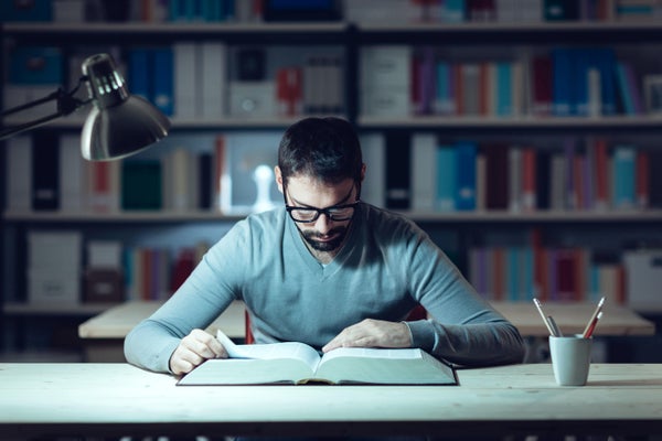 Young man in light blue sweater studying a book by a desk in front of a library of books.