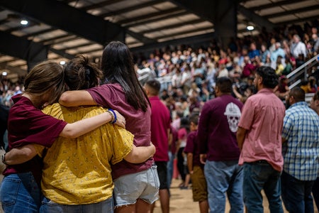 Three people in the foreground embrace each other while a crowd is viewed in background standing and in bleacher seats