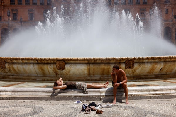 An exhausted woman lays in front of a fountain while her friend watches over her