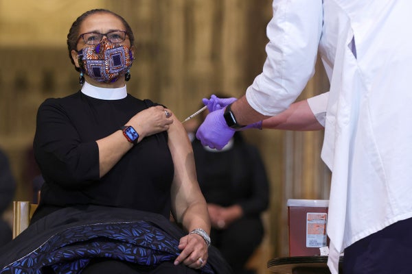 A masked woman wearing a clerical collar and glasses receives an injection.