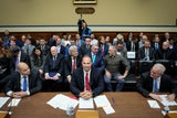 Three men sitting at a desk with people seated behind them in chairs