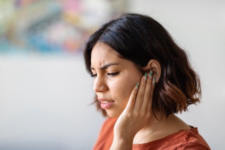 Portrait of a woman with dark hair and orange top touching her ear in pain.