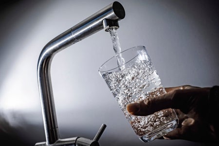 Person holding up a glass that's being filled by tap water against gray background.