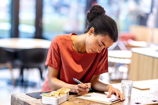 Woman with black hair and orange T-shirt writing in a coffee shop