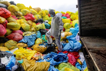 Worker in hazmat suit steps on massive mound of brightly colored trash bags.