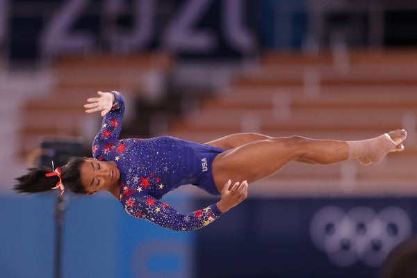 Simone Biles in a blue gymnastics costume with red stars appears to meditate in midair.