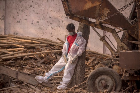 A man wearing a white jumpsuit and mask slumps against a tree amid wood scraps.