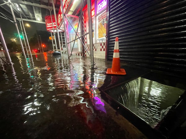 Store front sidewalk with orange traffic cone and basement latch flooding with water.