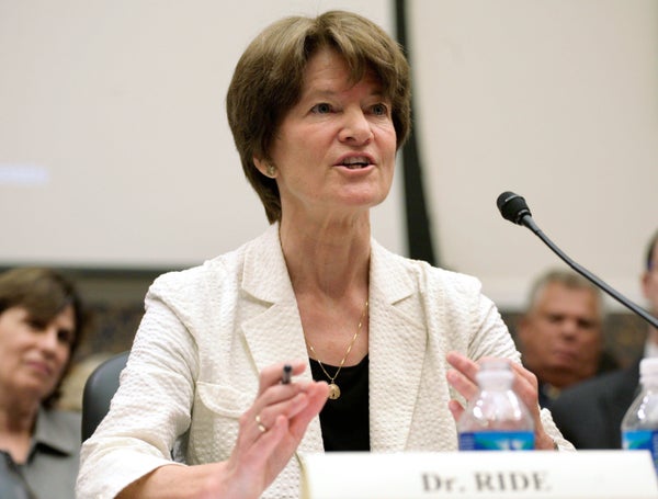 Sally Ride, in a white jacket, sits at a table in front of a microphone.