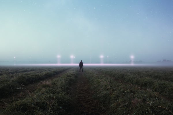 A man stands silhouetted in a field as mysterious lights glow in the background.
