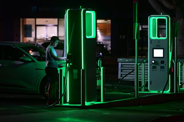 A driver charges his electric vehicle at night at a green neon lit charging station.