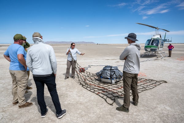 A team of researchers in the desert gather around a mock space capsule with a helicopter in background