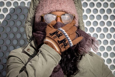 A rider braves freezing weather while waiting for a train in winter wearing full winter gear, holding up a scarf to cover their face and eye glasses completely fogged over