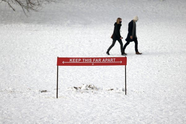 Couple walking across snowy park with COVID distancing sign in foreground.