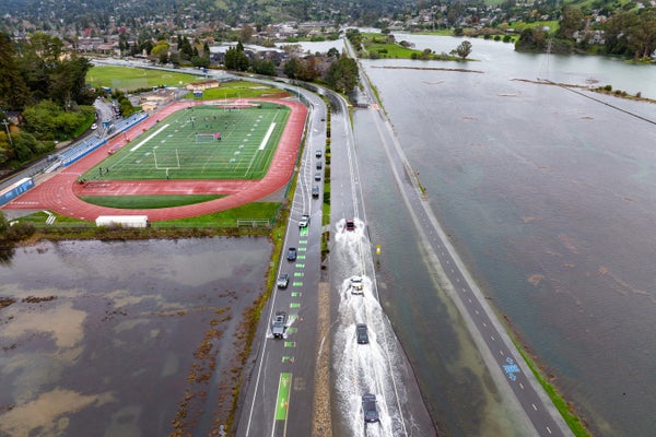 Vehicles driving along a road flooded with ocean water with a sports stadium on the left and homes and trees in the background.
