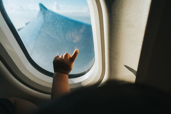 Baby's pointing hand shown in airplane window, interior view looking out the window with airplane wing outside.