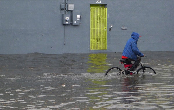 A cyclist with blue hoodie pedals through a flooded street with a yellow door on a building background.