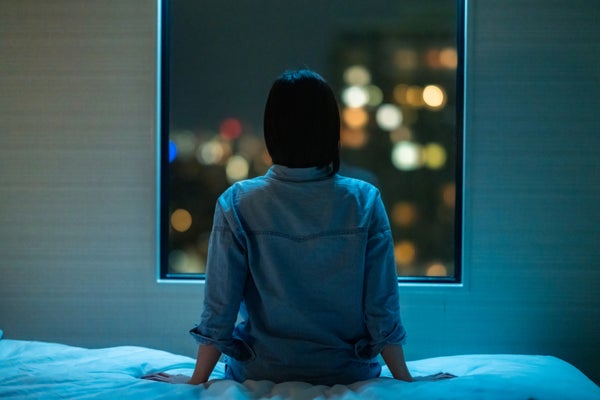 A woman sitting on the edge of a bed at night, shown from behind.