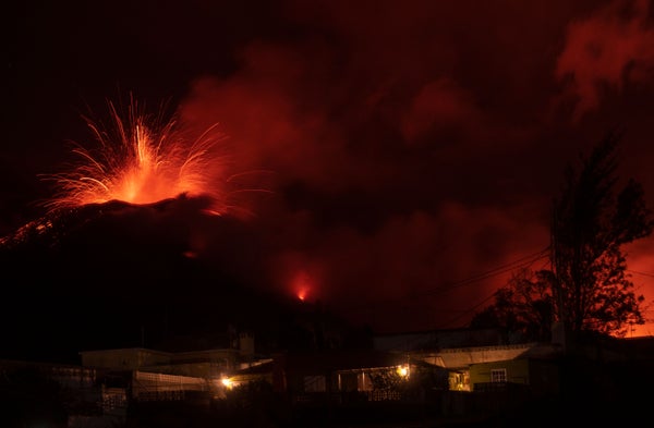 A volcano can be seen erupting in the distance as houses stand lit in the foreground.