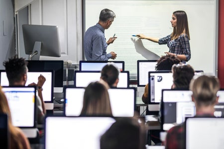 Professor and female student speaking in front of class of students using computers