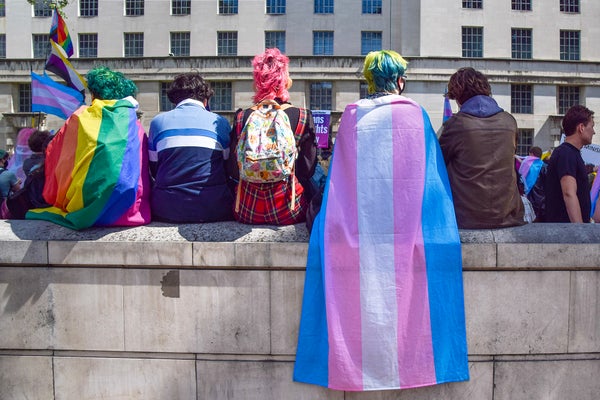 A group of five protestors sit on a stone wall, one wrapped in a pride flag, one wrapped in a trans tride flag, during a trans rights demonstration