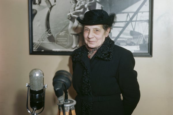 Lisa Meitner with black hat and coat in front of microphone