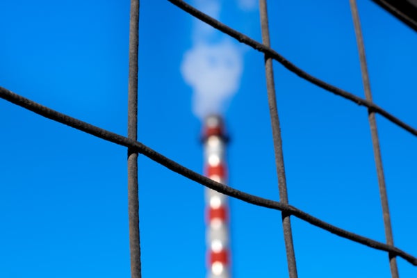 Grid metal fence and factory chimney releasing steam against the blue sky.
