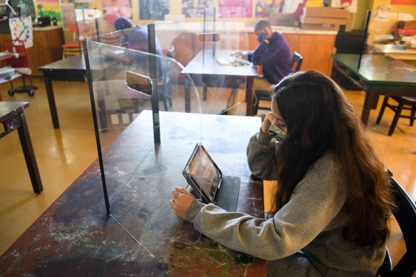 A girl sits at a desk in a classrom looking at a tablet, masked and behind a plastic barrier