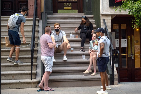 Group of five people having a conversation and drinking at the steps of an apartment building. One wears a mask.