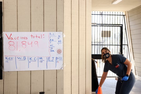 A woman with a mask stands next to a sign tabulating the administration of COVID vaccines.